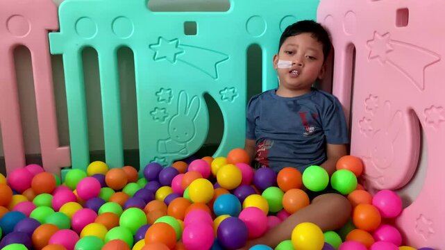 A child living with cerebral palsy is playing in a colorful ball pool in the occupational therapy room. A young child with a nasogastric tube sits among a colorful sea of plastic balls. 