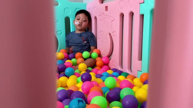 A child living with cerebral palsy is playing in a colorful ball pool in the occupational therapy room. A young child with a nasogastric tube sits among a colorful sea of plastic balls. 