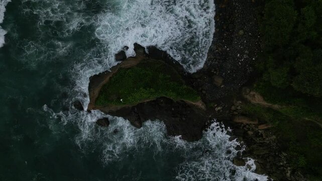 Aerial slow-motion footage of foamy sea waves crashing into rocks on Menganti Beach