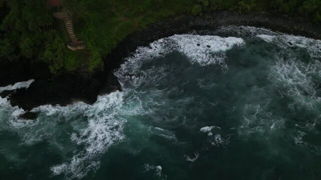 Aerial slow-motion of a rocky hill with foamy ocean waves on Menganti Beach in Kebumen, Indonesia