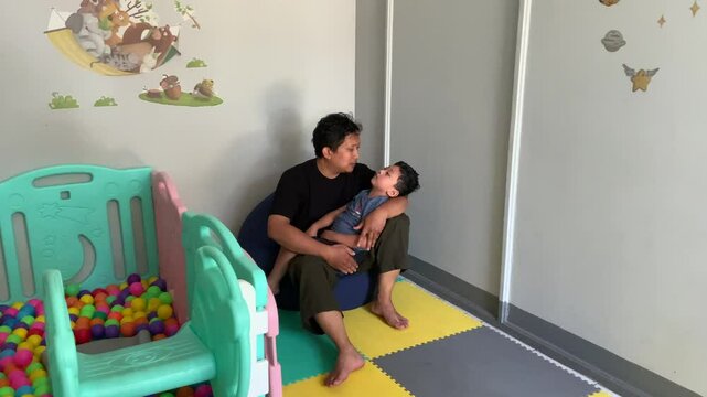 A father carries a child who lives with cerebral palsy and uses a nasogastric tube or NGT on his nose in the child's playroom and besides there is a colorful ball pool. The child looks sleepy.