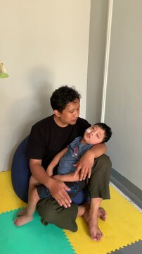 A father who is sitting holding a child who lives with cerebral palsy and wears a nasogastric tube or NGT on his nose in the child's playroom. The child looks sleepy