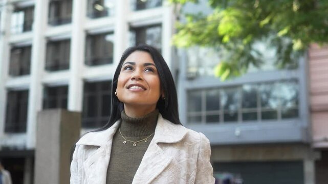 Optimistic professional woman in a stylish coat looking up at the city skyline with a thoughtful smile. Businesswoman admiring the urban architecture on a sunny day, feeling inspired