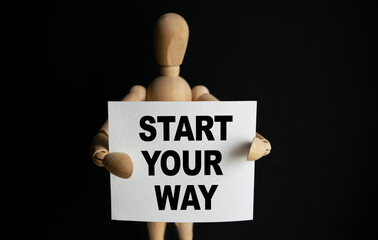 The wooden man holds a white sign with the text START YOUR WAY in his hands. The content of the lettering has implications for business concept and marketing.