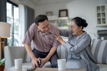 An elderly couple sits in their living room. The man holds his knee, appearing to be in discomfort, while his partner offers support. The image conveys care, concern, and the bond between the couple.