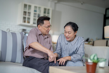 An elderly couple sits in their living room. The man holds his knee, appearing to be in discomfort, while his partner offers support. The image conveys care, concern, and the bond between the couple.