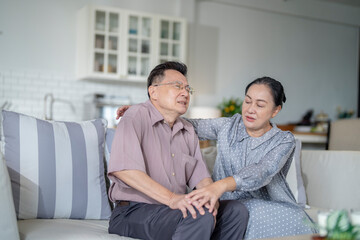 An elderly couple sits in their living room. The man holds his knee, appearing to be in discomfort, while his partner offers support. The image conveys care, concern, and the bond between the couple.