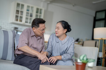 An elderly couple sits in their living room. The man holds his knee, appearing to be in discomfort, while his partner offers support. The image conveys care, concern, and the bond between the couple.