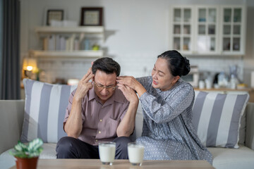 An elderly couple sits on a couch in a cozy living room. The man holds his head in discomfort, while his partner offers support. The image highlights care, concern, and the bond between the couple.