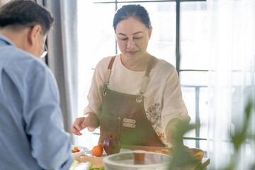 A woman prepares fresh vegetables in a cozy kitchen, while a man assists in the background. The scene emphasizes a warm and healthy lifestyle with a focus on fresh produce and home cooking.