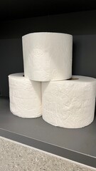 Vertical photo of three rolls of toilet paper neatly arranged on a bathroom shelf against a dark background. Minimalist composition highlighting hygiene and simplicity
