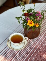 Vertical photo of a white porcelain cup of tea on a tablecloth beside a vase of flowers. Perfect for illustrating morning tea rituals, tea ceremonies, or cozy table settings.