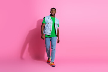 Trendy young man in casual denim vest walking confidently against a bright pink background.