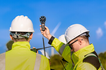 A team of engineers in high visibility safety gear stand together at a wind farm, conducting inspections and discussing renewable energy projects. Wind turbines surround them under a clear sky.