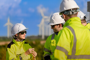 A team of engineers in high visibility safety gear stand together at a wind farm, conducting inspections and discussing renewable energy projects. Wind turbines surround them under a clear sky.