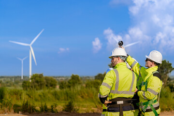 A team of engineers in high visibility safety gear stand together at a wind farm, conducting...
