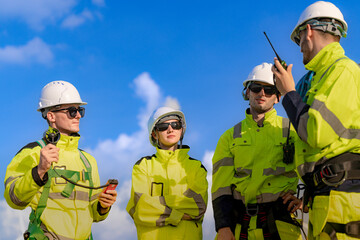 A team of engineers in high visibility safety gear stand together at a wind farm, conducting...