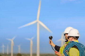 Two engineers wearing high visibility safety jackets and helmets measure wind speed with an...
