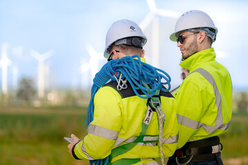 Two engineers in high visibility jackets are inspecting a wind farm. One is holding a mobile...