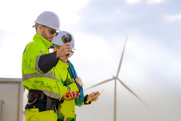 Two engineers wearing high visibility safety jackets and helmets measure wind speed with an...
