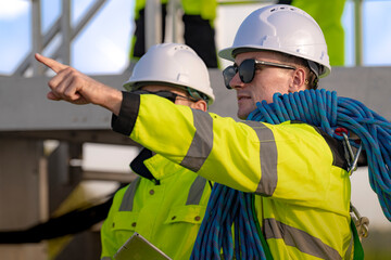 Fototapeta premium Two engineers in safety helmets and high visibility jackets engage in a field inspection. One is pointing while carrying a safety rope, highlighting communication, safety, and teamwork.