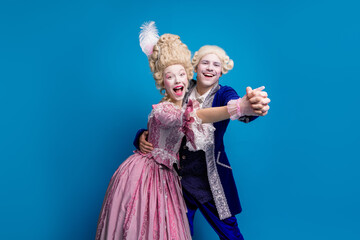 A joyful couple dressed in vintage aristocratic costumes shares a dance against a vibrant blue...