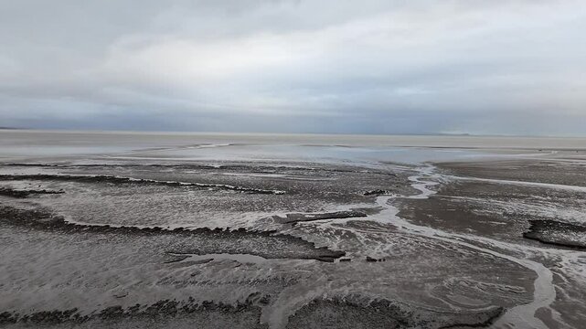 Drone glides low over tidal channels and mudflats at Morecambe Bay, revealing intricate patterns in the wet sand under a cloudy sky. Water carves through the landscape, creating reflective surfaces.