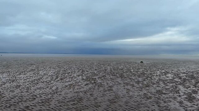 Fast and low drone flight over the wet rippled sands of Morecambe Bay under a cloudy sky. The waterlogged sand reflects the light as the horizon stretches across the coastal expanse.