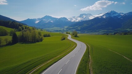 Naklejka premium A winding road through lush green fields and majestic mountains under a blue sky.