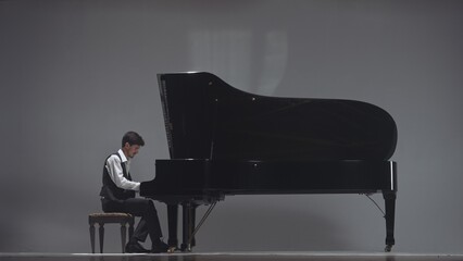 Pianist sitting and playing on the black grand piano on dark concert stage under bright spotlight, isolated on grey studio background. Artist performing in concert hall. © kinomaster