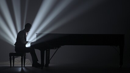 Pianist sitting and playing on the black grand piano on dark concert stage against bright...