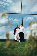 couple standing against the background of a large glass building. The woman is dressed in a white...