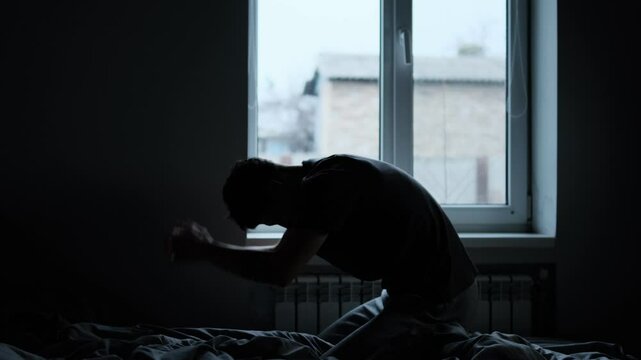Silhouette of a man sitting on a bed by the window, emotionally hitting the bed with fists, expressing deep despair and emotional struggle.