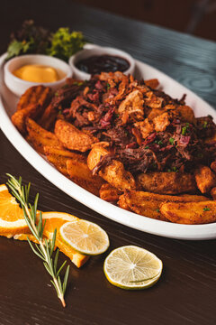 Chorrillana tradicional chilena con papas fritas r&uacute;sticas, carne mechada y salsas en un bar. Plato para compartir con c&oacute;ctel en mesa de madera.