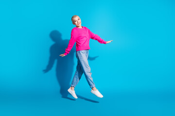 Joyful young woman jumping in mid-air against a vibrant blue background, wearing a bright pink sweater and jeans