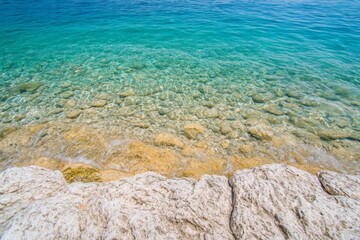 Obraz premium Serene Coastal Scene: Crystal Clear Water and Rocks at the Shore. Stunning Nature Photography of a Tranquil Beach. A Perfect Summer Day by the Ocean. A Peaceful and Beautiful View of the Shore.