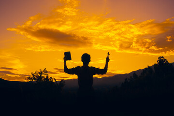 man standing praying and holding christian cross and bible for worshipping God at sunset background. christian silhouette concept.