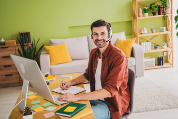 Cheerful man working remotely in a cozy home office environment with headphones, writing notes, and...