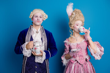 Elegant couple dressed in Baroque costumes drinking tea, set against a striking blue background