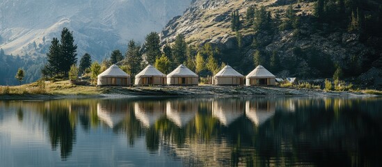 Fototapeta premium Yurts on the shore of a serene lake surrounded by majestic mountains reflecting in the water during a tranquil sunrise.