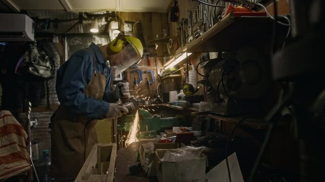 Medium full shot of male industrial worker in denim shirt, apron, gloves, protective earmuffs and face shield cutting metal on worktop in garage, with rain of spectacular flying sparks