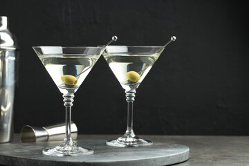 Martini cocktail with olives in glasses on grey table against black background