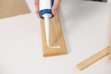 Man with caulking gun glueing wooden plank at white table, closeup