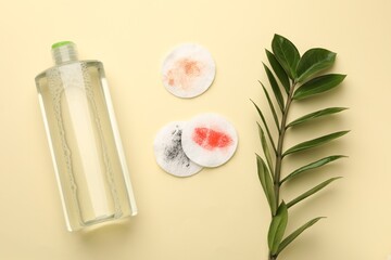 Dirty cotton pads, bottle of makeup remover and green branch on beige background, flat lay