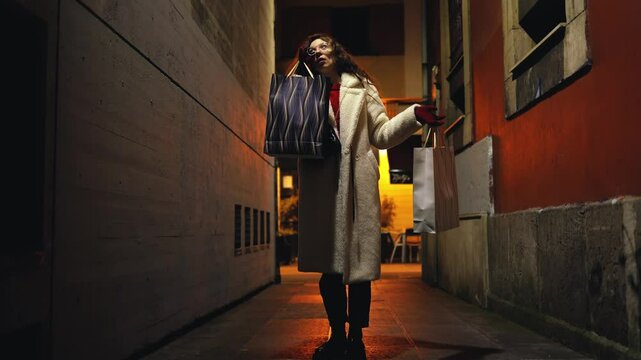 Happy curly girl runs from shopping with gift bags in her hands, impressionable, consumption, shops, shopping, dance, energy, movement, money, holidays, night, city alley, fashion, clothes, acting