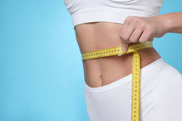 Weight loss. Woman measuring her slim waist with tape on light blue background, closeup. Space for...