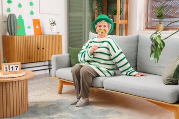 Senior woman in leprechaun hat with paper mustache sitting on sofa at home. St. Patrick's Day...