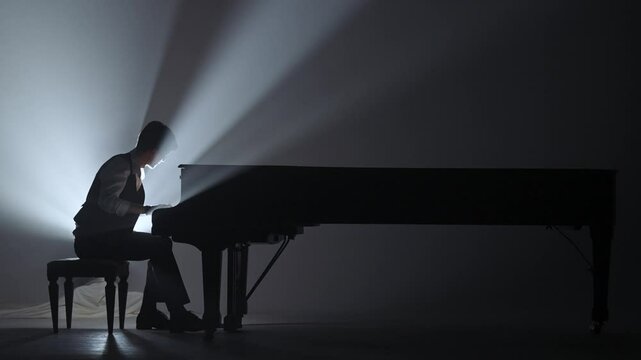 Pianist sitting and playing on the black grand piano on dark concert stage against bright spotlight, isolated on black studio background. Artist performing in concert hall.