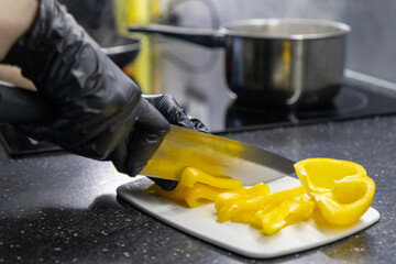 slicing yellow bell pepper with sharp knife on cutting board by gloved hand in kitchen, meal preparation process