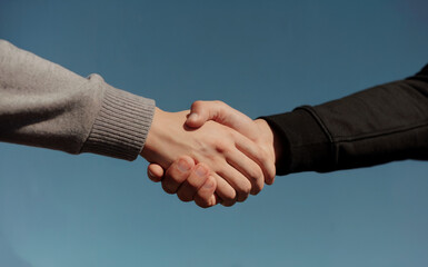 Two hands, one in a light gray sweater and the other in a black jacket, shaking hands against a clear blue sky. 